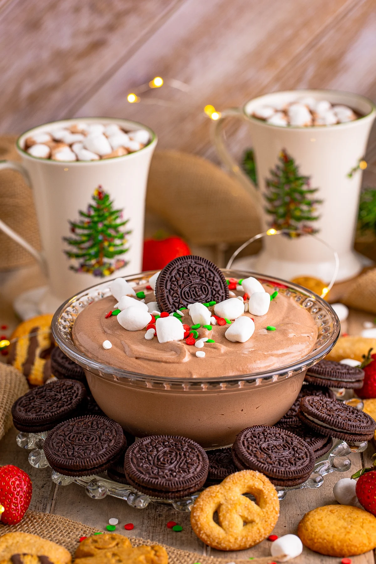 This is a photo of Hot Chocolate Dip served in a clear glass bowl topped with mini marshmallows, red, white and green sprinkles. The glass bowl in on a platter served with Oreos and butter cookies. 