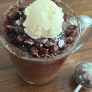 Close up of the Original Wacky Mug Cake topped with a scoop of vanilla ice cream, chocolate chips, and a dusting of powdered sugar in a clear glass mug.