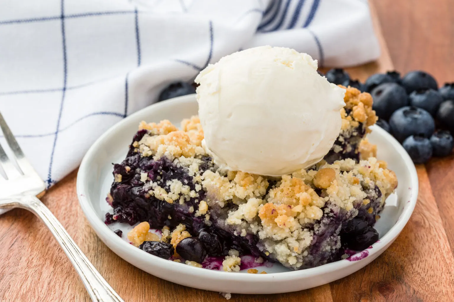 This photo shows blueberry crisp served in a white bowl with a scoop of vanilla ice cream on top. 