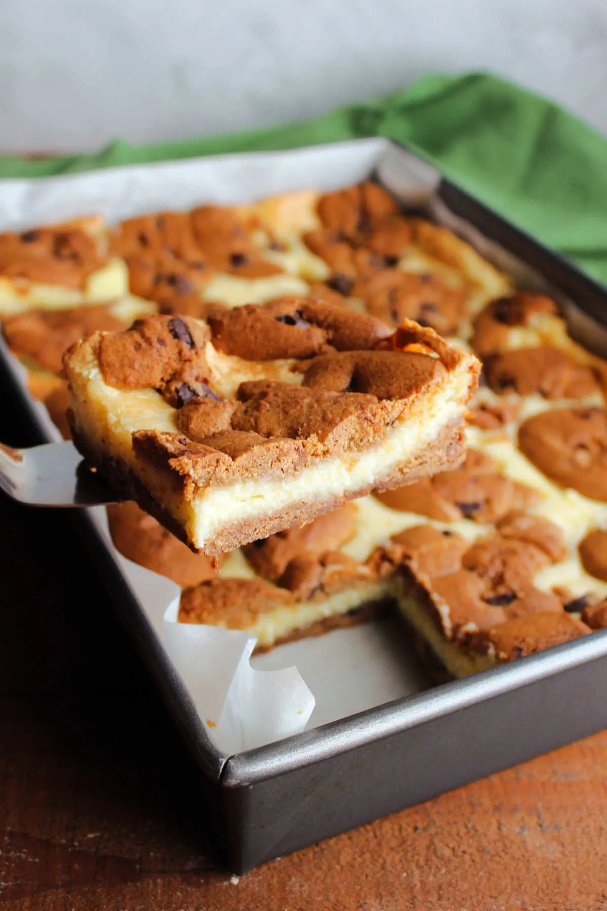 This photo shows Chocolate Chip Cookie Cheesecake Bars in a baking pan after baking with a spatula holding a bar, ready to serve. 