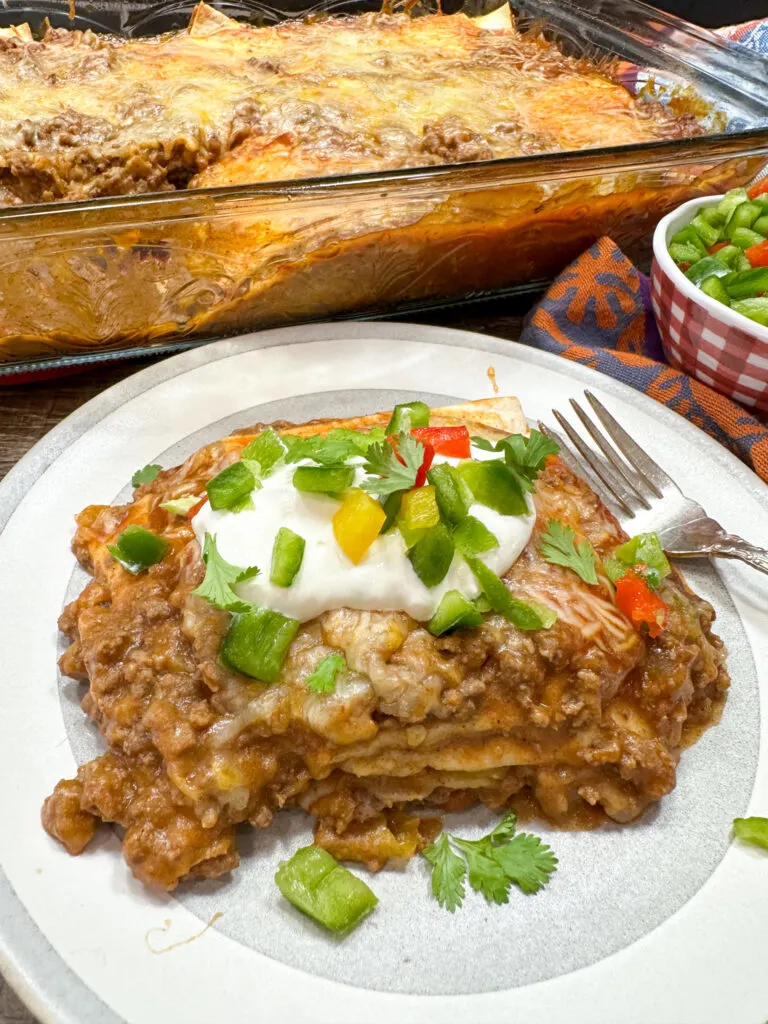 This photo shows a serving of beef enchilada casserole on a plate. Behind the plate is the baking dish with the casserole. 
