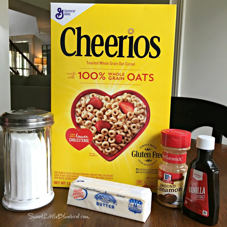 This is a photo of a box of Cheerios, stick of butter, sugar, vanilla extract and ground cinnamon on a kitchen table.