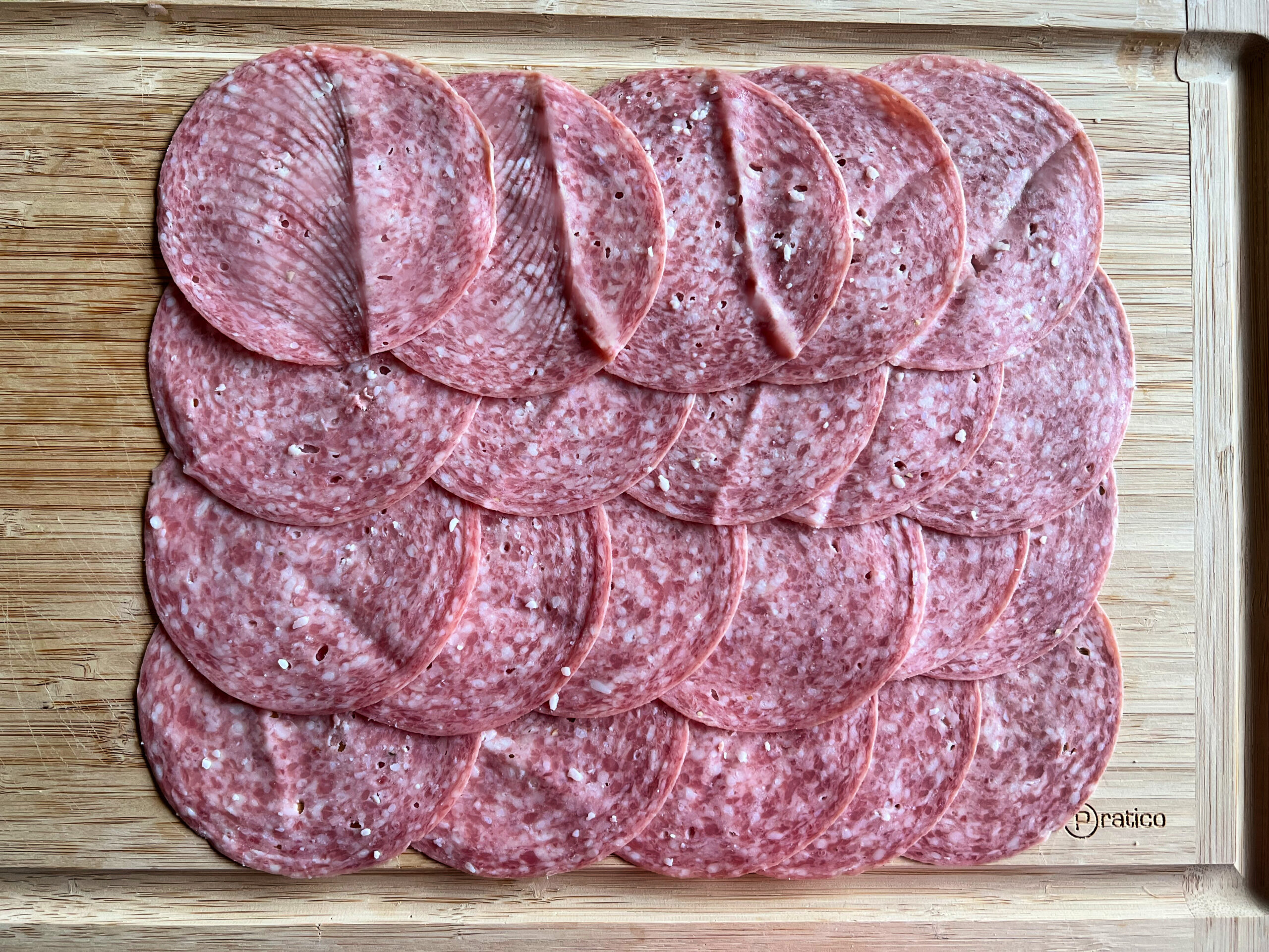 This photo shows salami slices on a cutting board placed in 4 rows, with 5 pieces of salami in each, overlapping, forming a rectangle. 