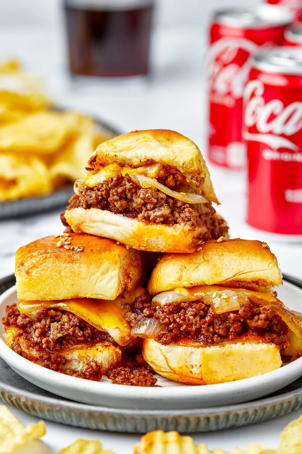 This photo shows 3 Sloppy Joe Sliders stacked on a plate. The Sliders are topped with melted cheddar cheese, served with potato chips and cans of Coke.