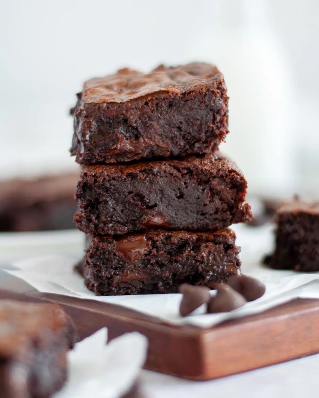 Easy Homemade Brownies Three brownie squares stacked on top of each other on a cutting board with parchment paper with a few chocolate chips next to them.